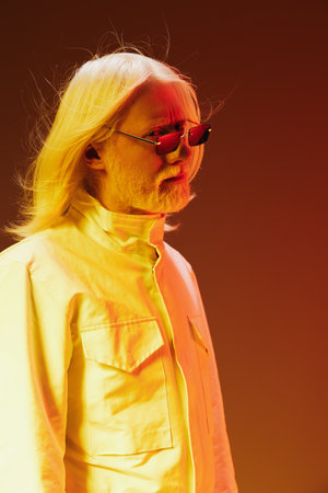 Stylish person with long blonde hair in sunglasses, wearing a white jacket, against a warm gradient...