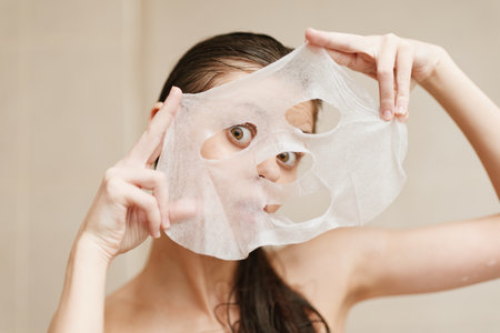 Surprised young woman holding a sheet mask up to her face while preparing for a skincare routine. Self-care and beauty concept with a playful expression.の写真素材