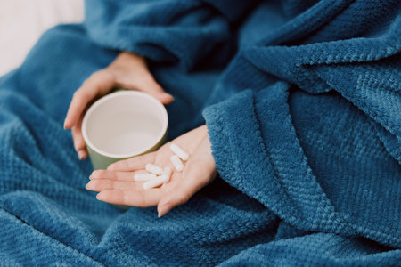 Person wrapped in blue blanket holding cup of coffee and pills in one hand, pill bottle in the otherの写真素材