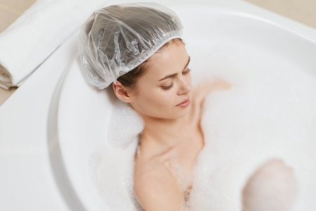 Serene young woman relaxing in a bubble bath, wearing a shower cap, enjoying self-care time in a cozy bathroom setting. Wellness and relaxation concept.の写真素材