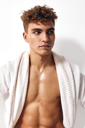 Stylish young man with curly hair wearing a white bathrobe, showcasing confidence and a relaxed attitude against a plain white background. Self-care and grooming concept.の写真素材