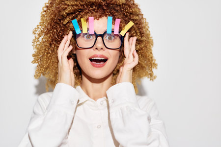 Funny woman with curly hair and colorful clips on glasses, expressing surprise against a plain white...