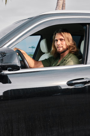 A man with long hair is sitting in a black car, looking out from the driver s seat against a...