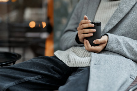 Fashionable man sitting in a chair with a cell phone, wearing a sweater and holding a cup of coffeeの写真素材