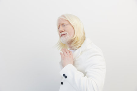 Portrait of a thoughtful man with long white hair and a beard, wearing a white coat against a minimalistic white background, exuding elegance and serenityの写真素材