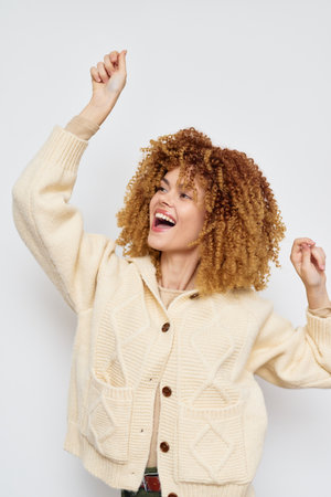 joyful woman dancing with curly hair in a stylish cream cardigan, joyful expression, bright background, showcasing fashion and happiness, perfect for lifestyle imageryの写真素材