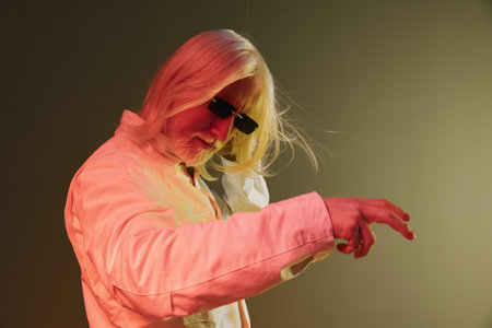 Stylish man with long blonde hair wearing sunglasses and a white jacket, posing dramatically against...