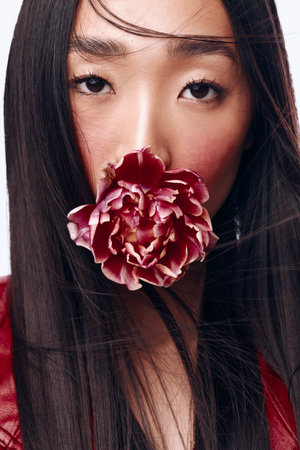 Beautiful woman with long black hair holding a flower in front of her face in red jacketの写真素材