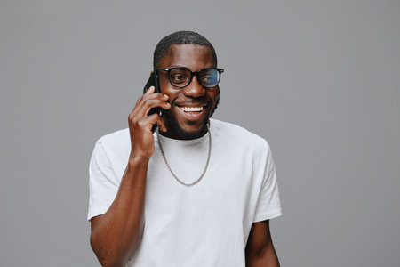 Smiling man talking on phone, wearing glasses, casual white t shirt, joyful expression, gray background, modern concept, communication, happiness evident in his demeanorの写真素材