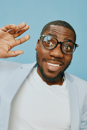 Smiling young man in glasses, wearing a light blazer, waving hello against a blue background,...