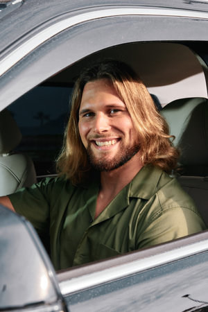 Smiling man with long hair in a car interior, wearing a green shirt, showcasing a friendly demeanor...