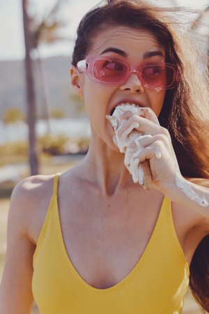 Woman enjoying ice cream outdoors in a sunny setting, wearing pink sunglasses and a yellow swimsuitの写真素材