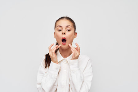 surprised woman showing shock or disbelief with hands on her face in casual attire, isolated on a light gray background, conveying emotions of amazement and astonishmentの写真素材