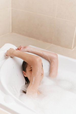 Relaxed young woman enjoying a bubble bath at home with arms resting on the tub, creating a peaceful...