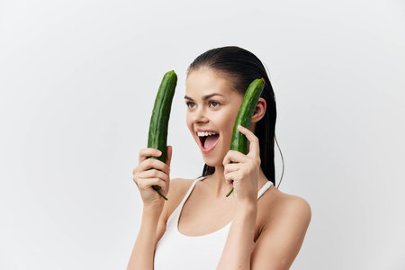 Woman holding cucumbers, playful expression, on white background, fresh and vibrant concept, healthy lifestyle, wellness, natural beauty, smiling and engagingの写真素材