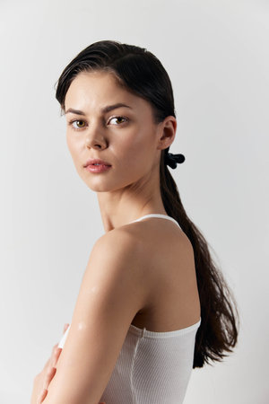 Portrait of a young woman with long dark hair and a ponytail, wearing a white sleeveless top, against a minimalistic light grey background, conveying beauty and simplicityの写真素材