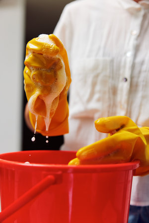 A woman in a white shirt and yellow rubber gloves is cleaning a bucket of soapy water with a spongeの写真素材