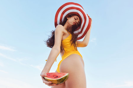 Young woman in a yellow swimsuit holding watermelon, wearing a large striped hat against a blue skyの写真素材