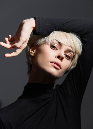 Stylish young woman with short blonde hair posing confidently against a dark background. Fashion and...
