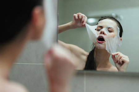 Young woman applying facial mask in a bathroom mirror, expression of surprise while removing the mask, promoting self-care and beauty rituals.の写真素材