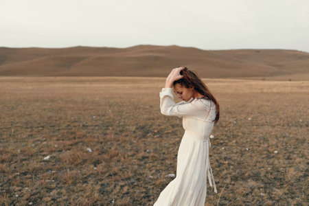 Lonely woman in white dress standing in sprawling field with arms raised in solitude and reflectionの写真素材
