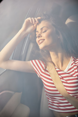 Young woman smiling while leaning against a car window, enjoying a joyful moment on a sunny dayの写真素材