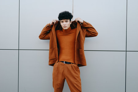 A young man in a stylish brown suit with a turtleneck poses confidently against a modern gray wallの写真素材