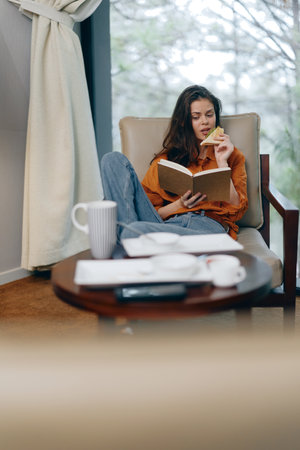 Cozy woman reading a book while enjoying a cookie and coffee in a tranquil indoor setting, wearing casual attire with an inviting atmosphereの写真素材