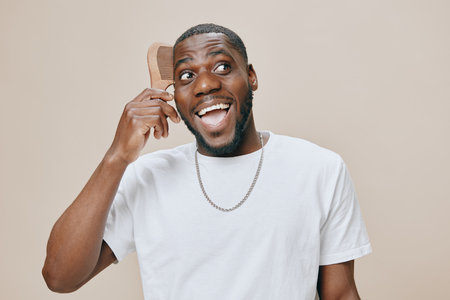 Happy man grooming with a comb, smiling joyfully against a soft beige background, wearing a casual white t shirt, showcasing a vibrant and carefree spiritの写真素材
