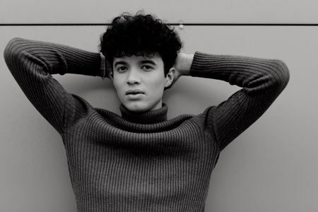 Fashionable black and white portrait of a young man leaning against a wall with hands on head
