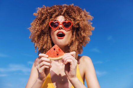 Young woman with curly hair wearing heart shaped sunglasses, holding a slice of watermelon on a sunny beach dayの写真素材