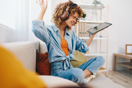 Smiling Woman Holding Tablet PC, Enjoying Music and Chatting Online on Cozy Couch in Living Roomの写真素材