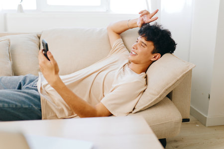 Relaxed young man using smartphone on a cozy sofa, comfortable living space with neutral colors The vibe is casual and inviting, perfect for lifestyle imageryの写真素材