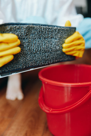 Person in rubber gloves holding a sponge next to a bucket with a red lid on a wooden floorの写真素材