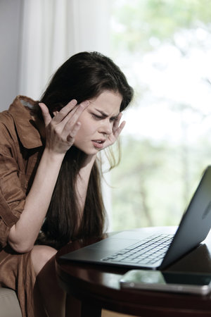 Young woman expressing stress while working on a laptop in a cozy home environment, featuring a brown outfit and natural backgroundの写真素材