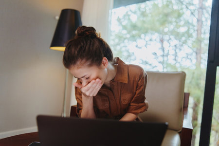 A young woman in a brown shirt sitting in a modern living room, covering her mouth with laughter while looking at a laptop, conveying joy and relaxationの写真素材