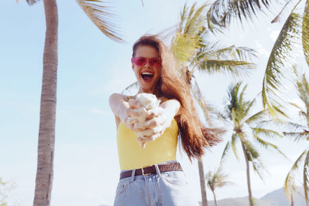 Young woman in yellow top enjoying summer under palm trees, smiling with a playful expression, surrounded by bright sunlightの写真素材