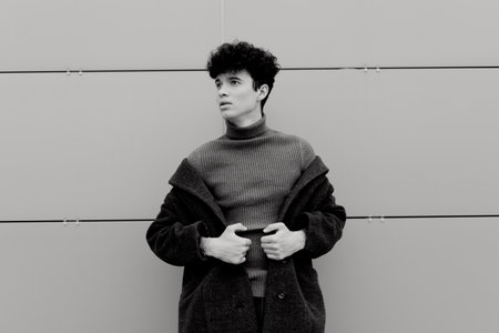 Fashionable young man in a sweater and coat, standing in front of a wall with hands in pockets in...