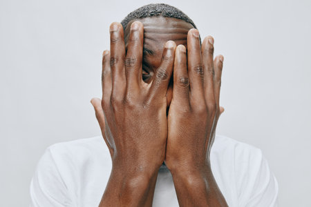 Emotional man hiding face with hands, expressing vulnerability and introspection, wearing a simple white shirt against a neutral light gray backgroundの写真素材