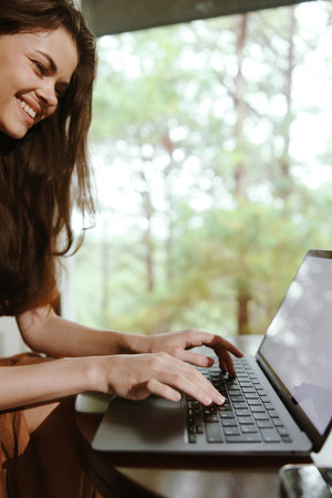 Young woman in casual attire, smiling while working on her laptop in a serene, nature inspired office setting with greenery in the backgroundの写真素材