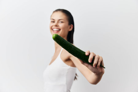 Smiling woman holding a cucumber in a playful manner The background is light and neutral, emphasizing her joyful expression and the vibrant green of the cucumberの写真素材