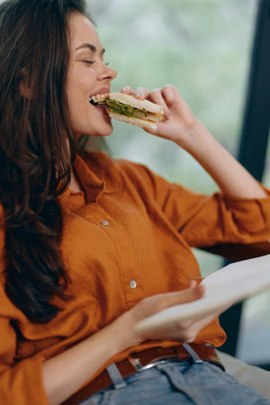 Young woman enjoying a delicious sandwich in a cozy indoor setting, wearing a stylish orange shirt, showcasing happiness and relaxation with fresh ingredientsの写真素材