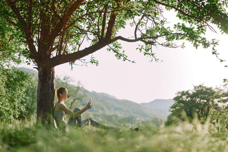 Young woman enjoying a refreshing drink under a tree in a serene natural setting, emphasizing relaxation, summer vibes, and healthy lifestyle choicesの写真素材