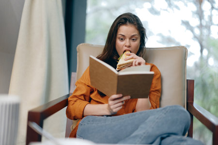 Young woman enjoying a sandwich while reading a book, embodying a relaxed and cozy atmosphere with...