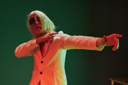Stylish man in white suit with futuristic sunglasses striking a dynamic pose against a dark green background, illuminated with vibrant red and green lighting for a striking visual effectの写真素材