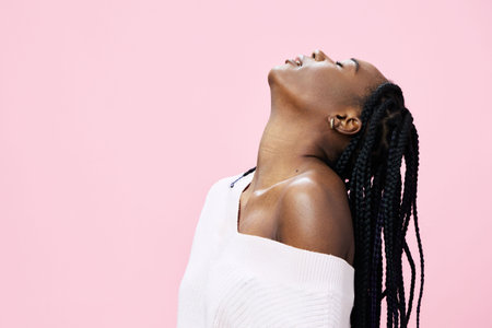 Young Black woman with braided hair, wearing a white sweater, exudes joy against a pastel pink background, perfect for fashion and emotional wellness themesの写真素材