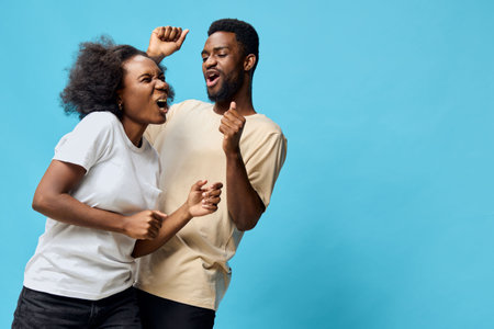 A joyful young African couple dancing together against a vibrant blue background, expressing...
