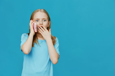 Surprised girl talking on smartphone with blue background, expressing shock and curiosity, dressed in casual light blue t shirtの写真素材