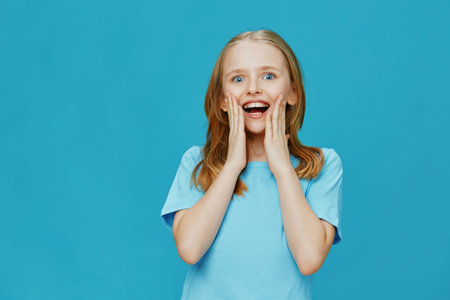 Happy girl in a blue shirt expressing excitement against a bright blue background, showcasing joy and surprise in a vibrant settingの写真素材