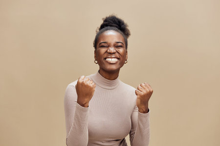 Happy African woman celebrating success in a light beige background, wearing a stylish turtleneck and expressing joy and excitement with clenched fistsの写真素材